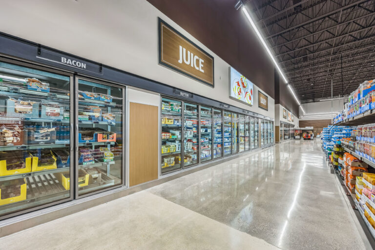 A grocery store aisle featuring long rows of refrigerated glass-door cabinets labeled for bacon, juice, butter, yogurt, and dairy.