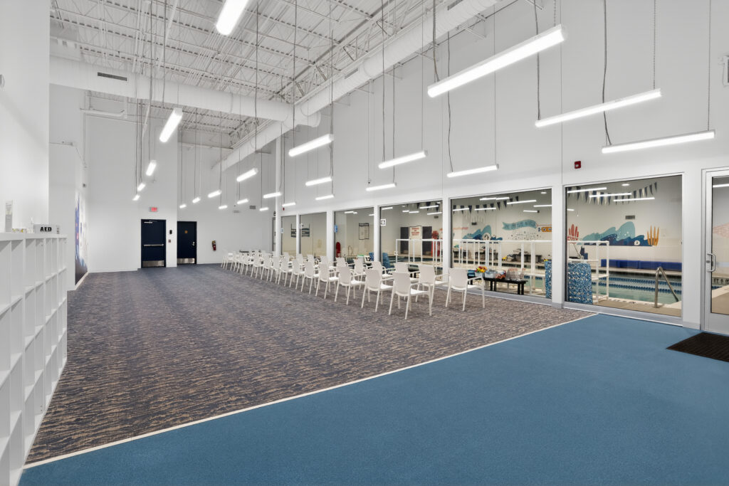 A row of white chairs lines a viewing window overlooking an indoor swimming pool.