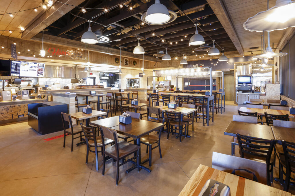 Dining area of a casual restaurant with wooden tables, chairs, and an open kitchen counter in the background.