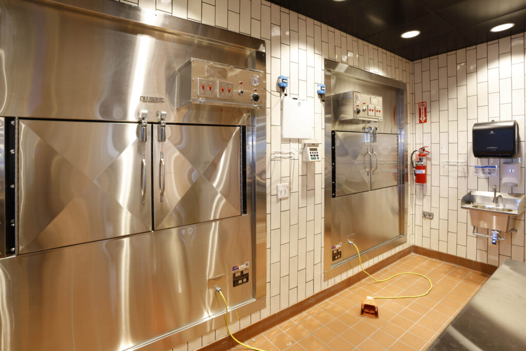 Two industrial stainless steel ovens installed in a white tiled wall, with a hand-washing station nearby.