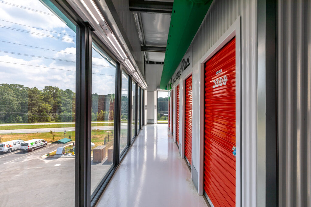 A hallway inside a storage facility with a row of red roll-up doors on the right and a glass wall on the left overlooking a parking lot.