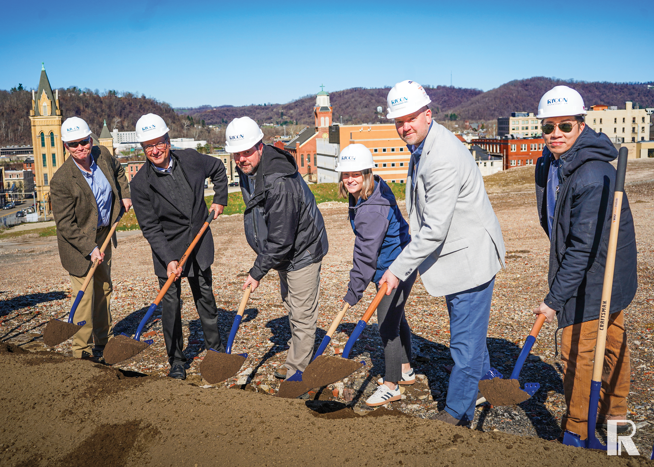 Six people in hard hats participate in a groundbreaking ceremony, each holding a shovel in a pile of dirt with a town skyline in the background.