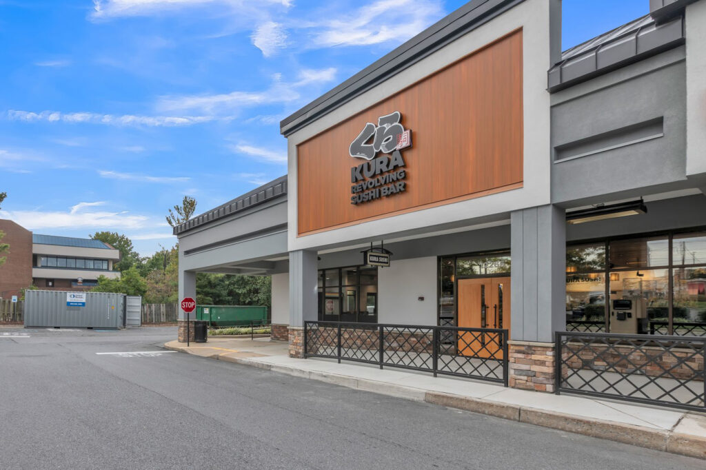 Exterior of a Kura Revolving Sushi Bar restaurant with a wooden facade and a black metal railing in front of the entrance.