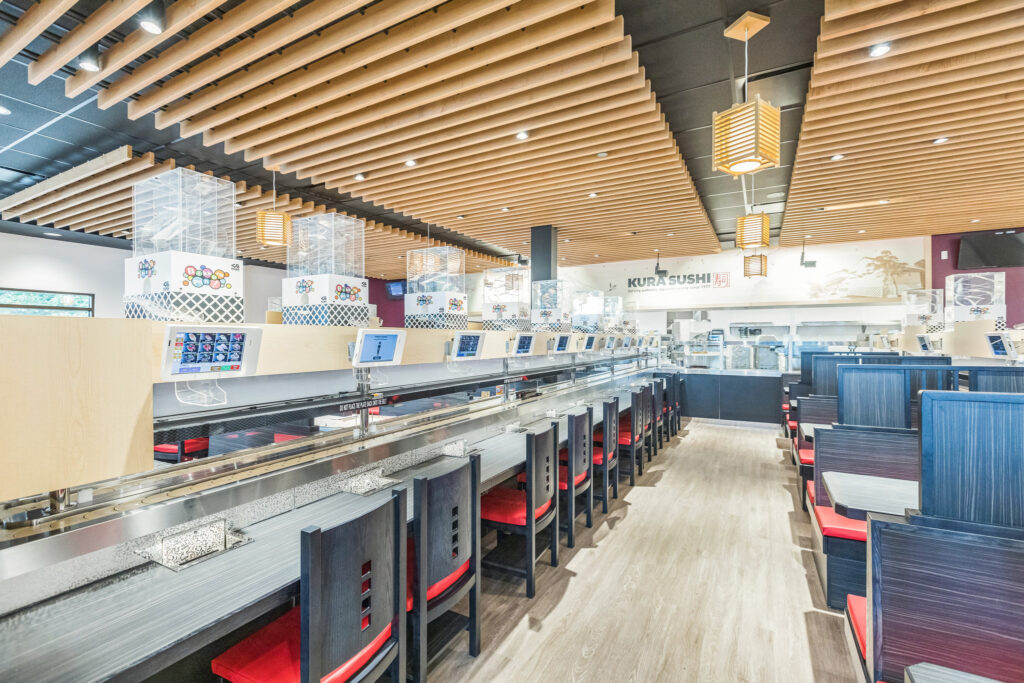 Interior of a Kura Sushi restaurant featuring a conveyor belt system, individual booths with tablet ordering screens, and wood-slat ceiling accents.
