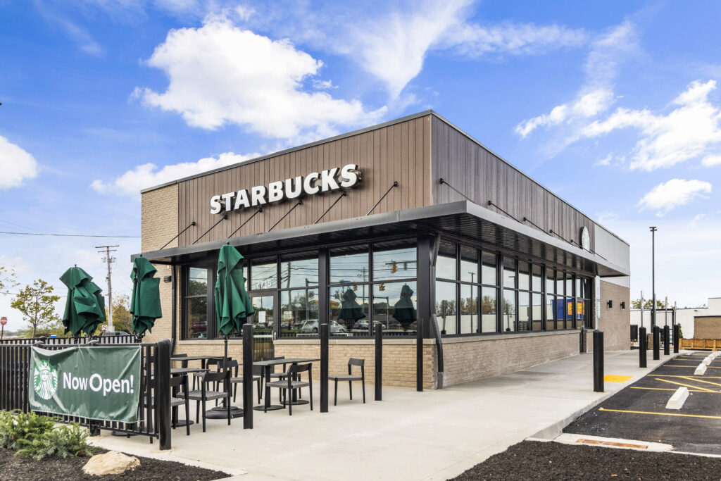 Exterior view of a Starbucks with an outdoor seating area and a “Now Open!” sign.