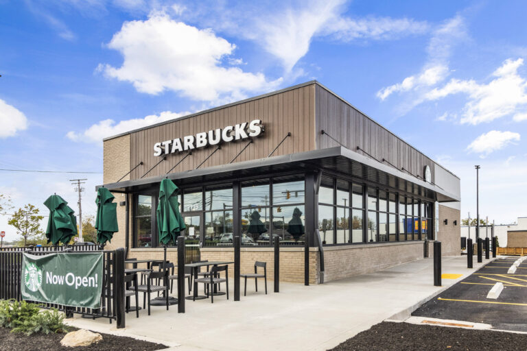 Exterior view of a Starbucks with an outdoor seating area and a “Now Open!” sign.