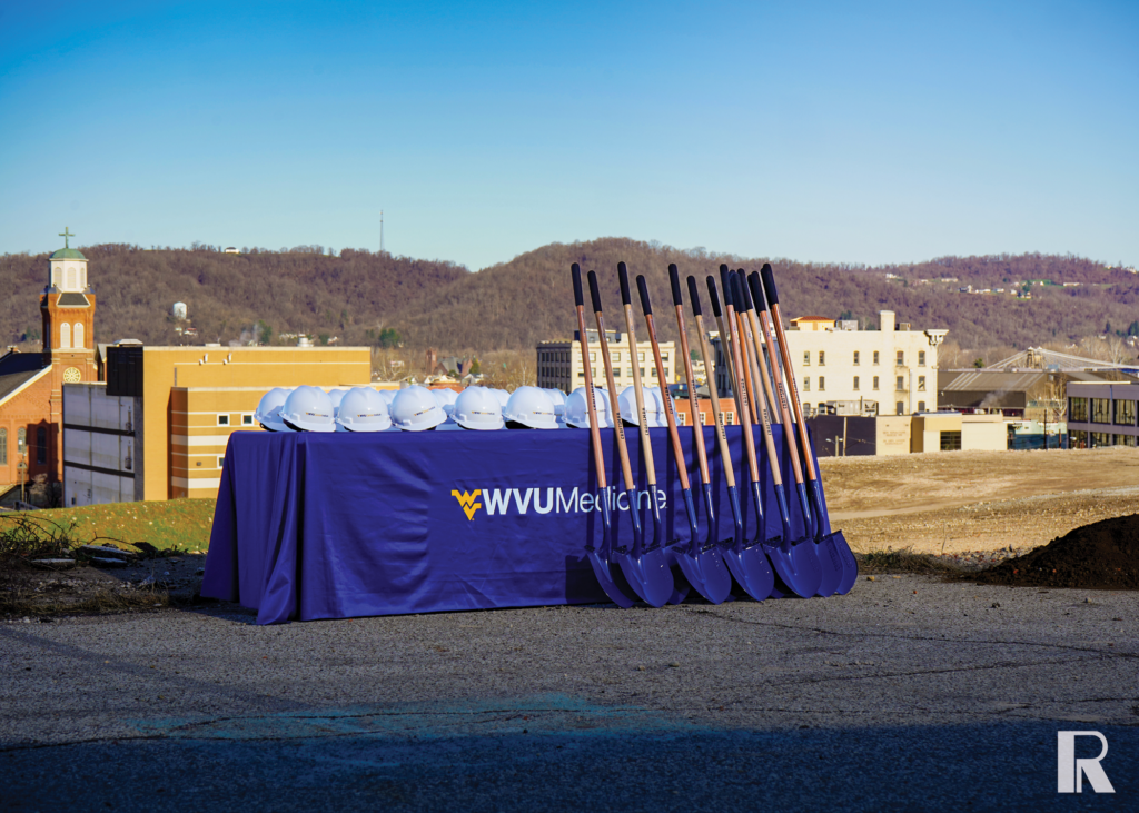 A table draped in a blue WVU Medicine cloth holds several white hard hats and a row of ceremonial shovels at a construction site.