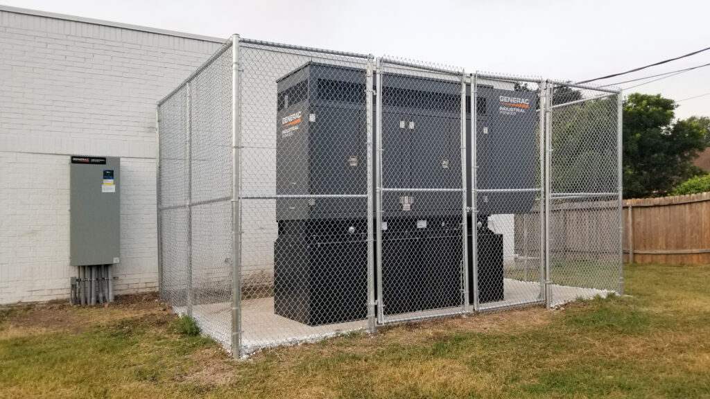A large Generac industrial power generator housed inside a chain-link fence enclosure next to a building.