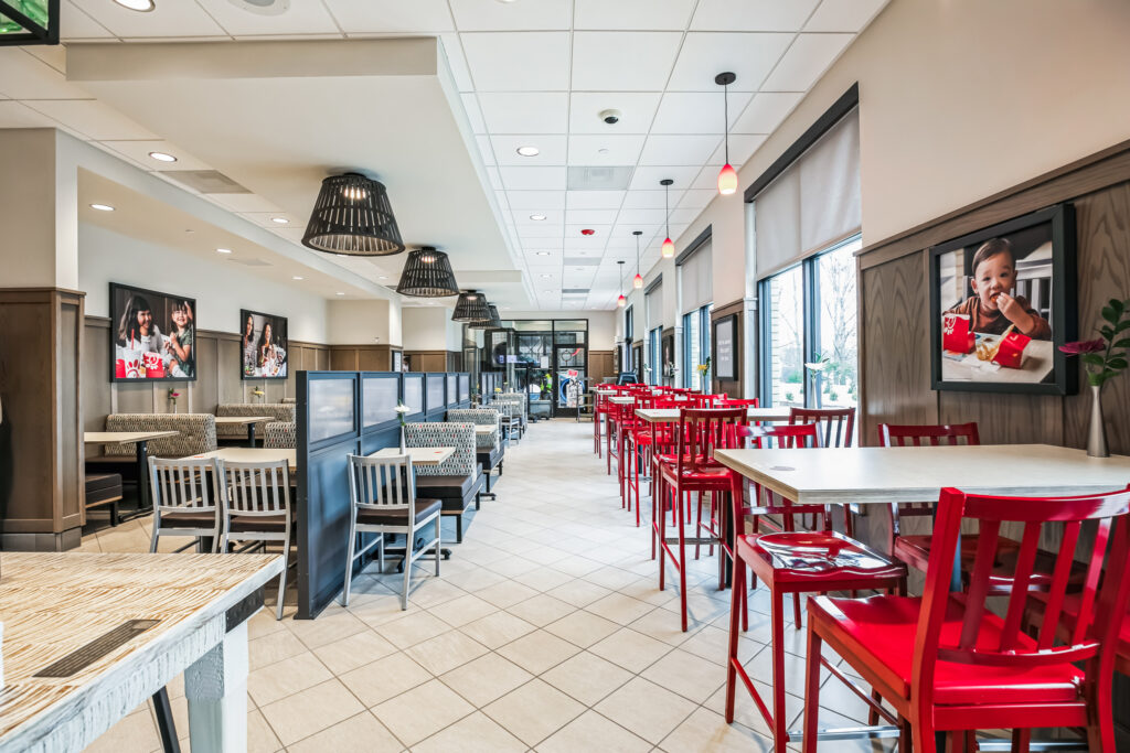 Interior of a brightly lit restaurant with white tile floors, tables, and rows of red chairs.