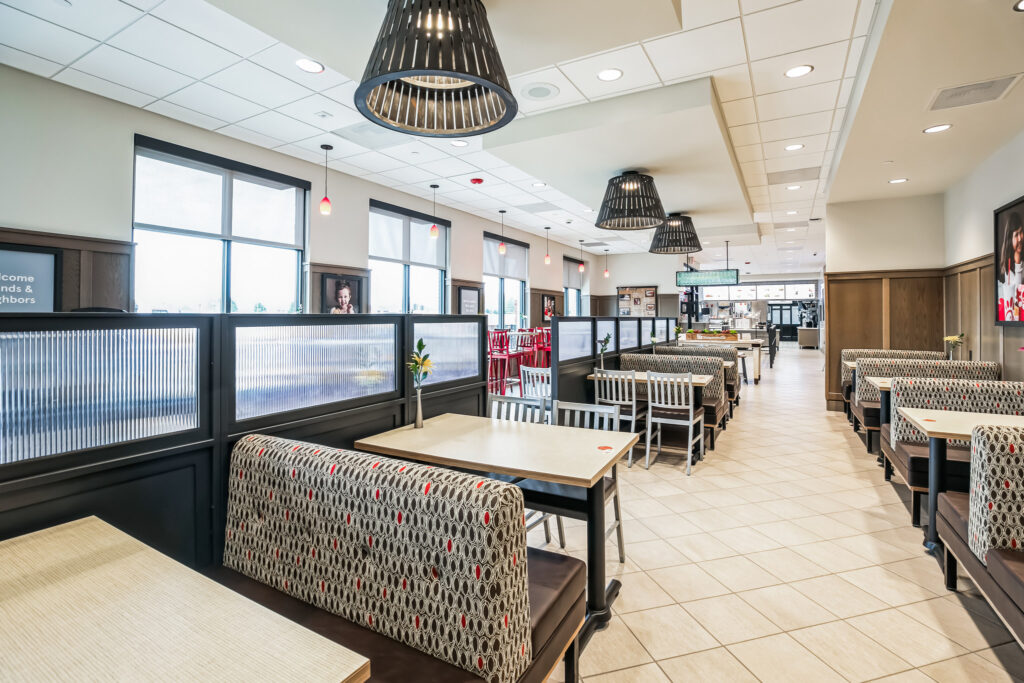 Interior of a restaurant dining area with booths featuring patterned upholstery, wooden tables, and dark pendant lights hanging from a drop ceiling.