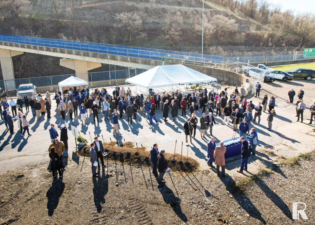 A large group of people gathers at a construction site for a ceremonial groundbreaking event with shovels lined up in the dirt.