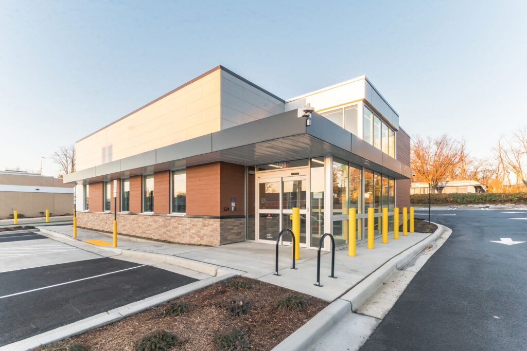 Exterior view of a modern commercial building with a glass entrance, brick and metal siding, and yellow bollards.