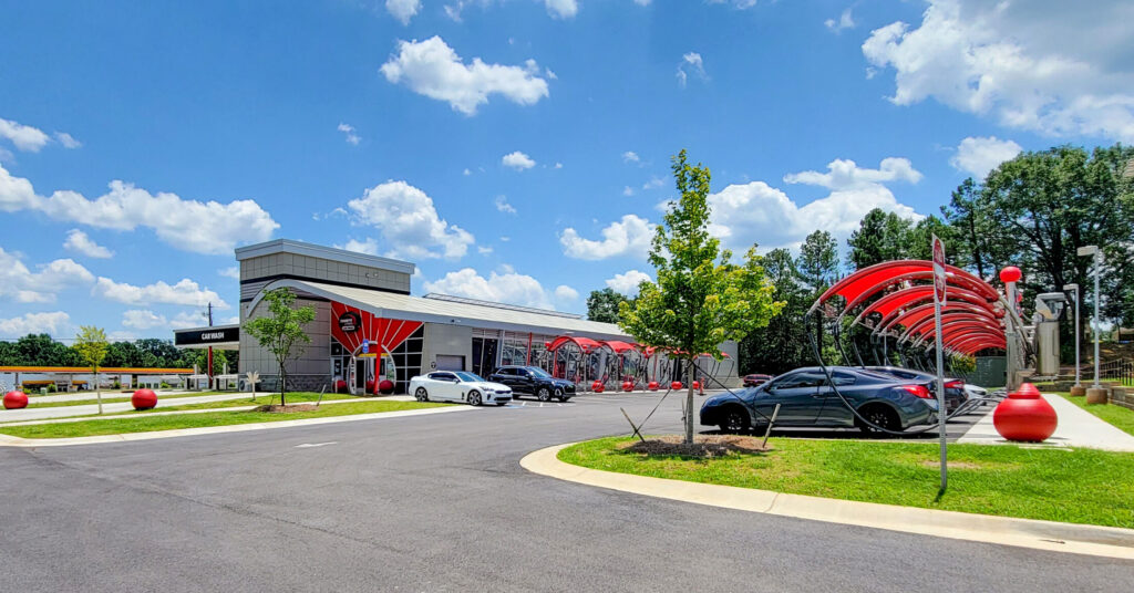 A car wash facility with a drive-thru lane, several parked cars, and red archway structures under a bright blue sky.