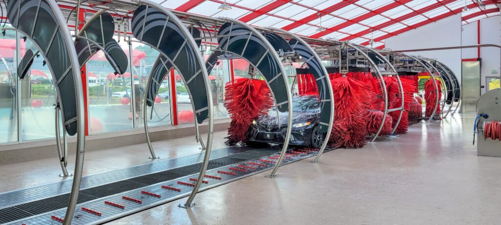 A car moving through an automated car wash with large, circular metal arches and red rotating brushes.