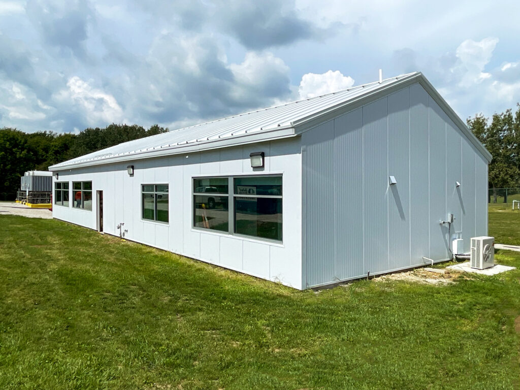 A single-story, rectangular white metal building with multiple windows situated on a grass lot under a cloudy sky.
