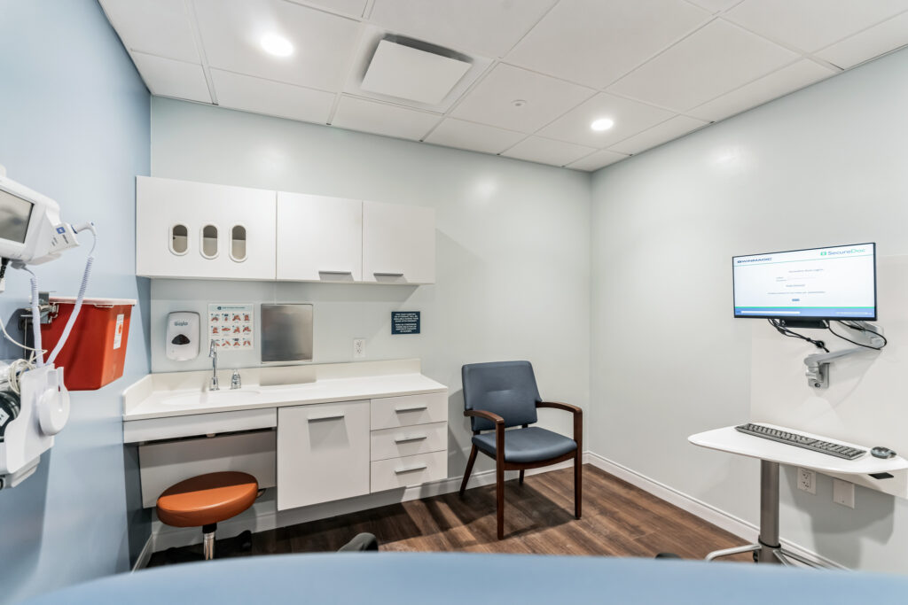 An examination room with a sink, white cabinetry, a blue chair, and a wall-mounted computer screen showing a login page.