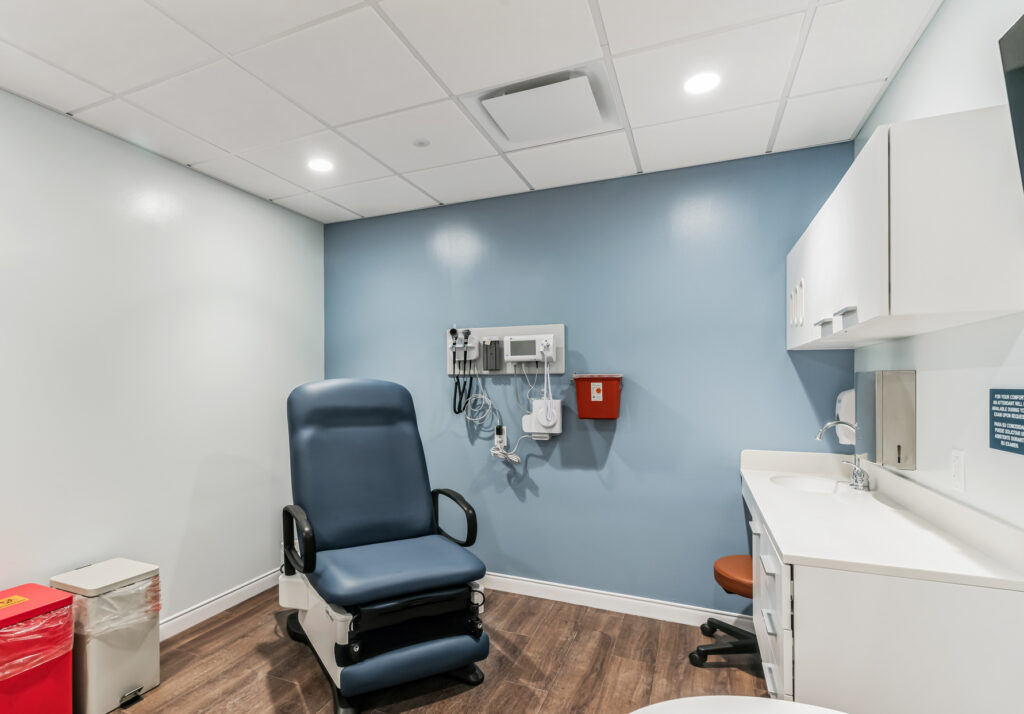 A medical examination room with a blue exam chair, wall-mounted diagnostic tools, a sink, and cabinetry.