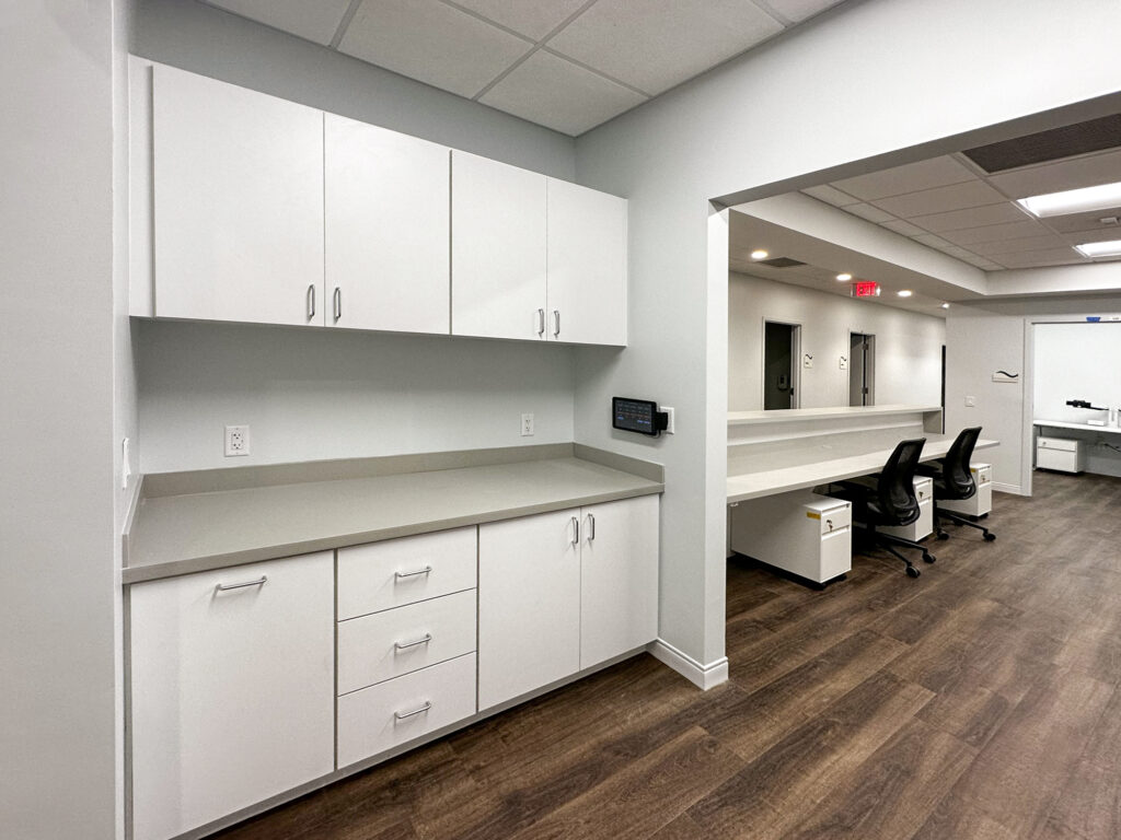 Office kitchenette with white cabinets and a gray countertop, leading to a hallway with desks and rolling chairs.