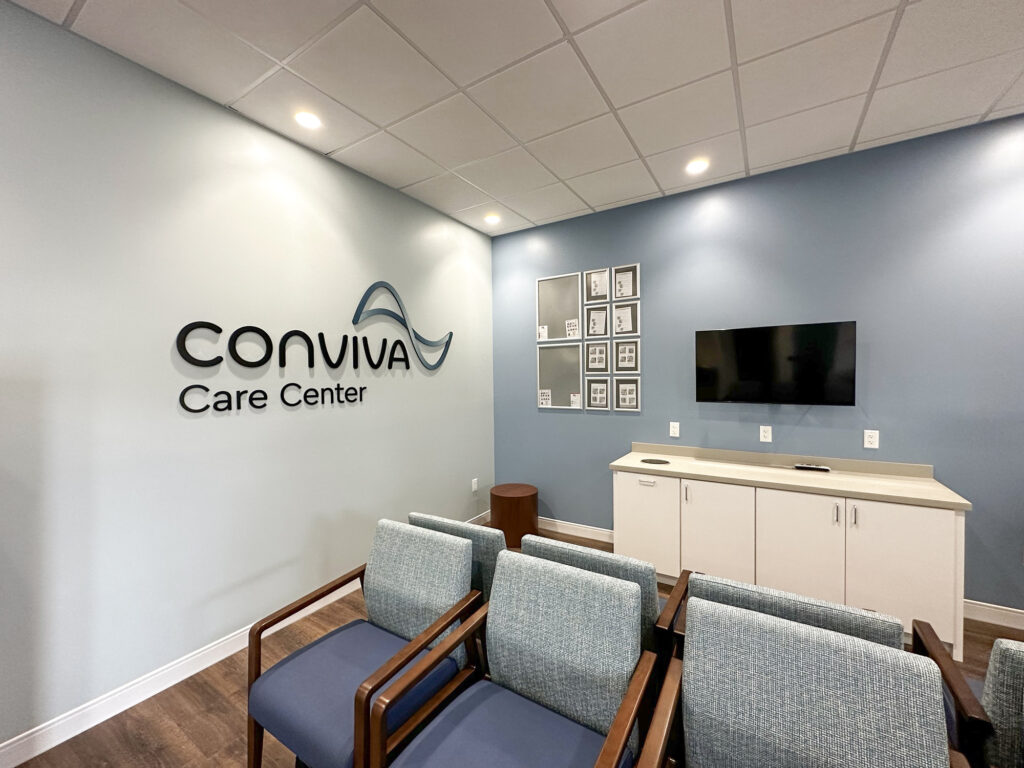 A Conviva Care Center waiting room with rows of chairs, a television, and a grid of framed photos on the wall.