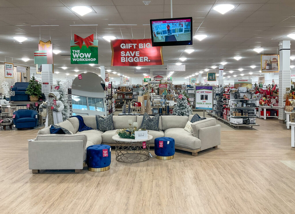 A beige sectional sofa with blue accent pillows and two matching blue ottomans in a furniture store aisle, with seasonal holiday decorations in the background.