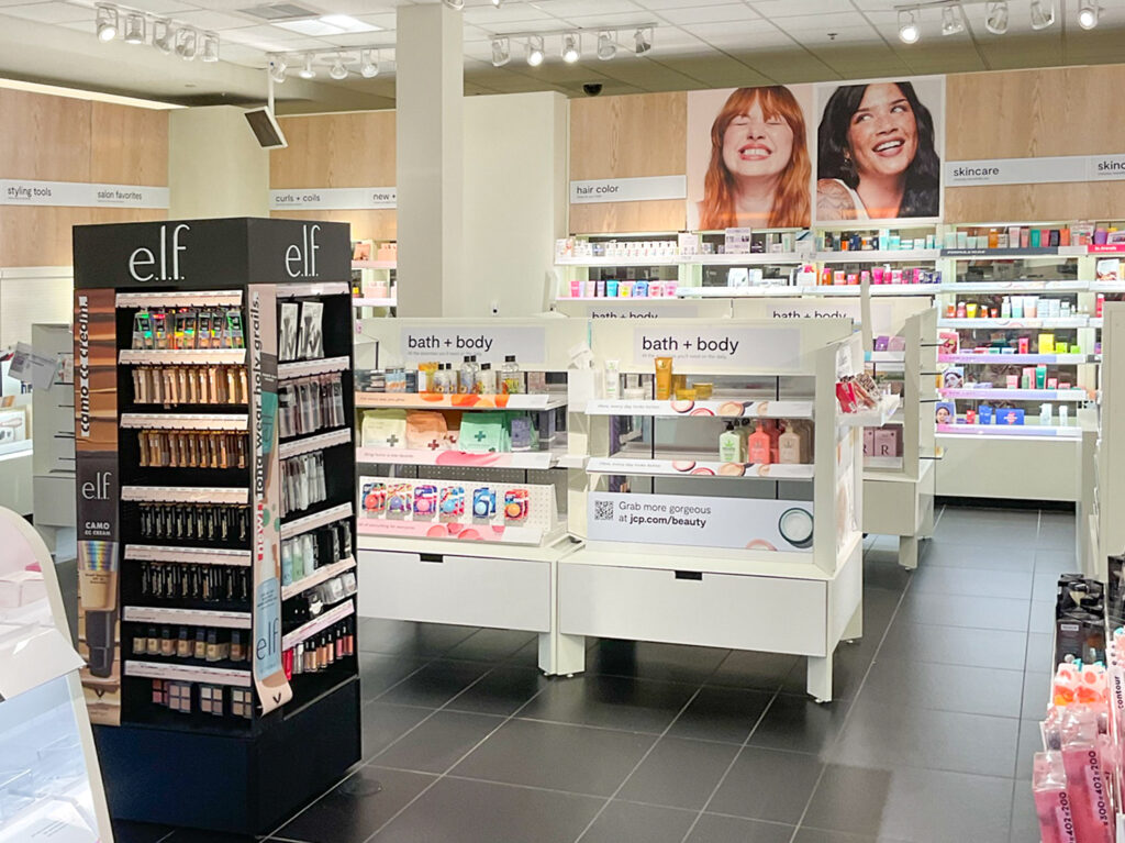 Inside a beauty retail store, a black e.l.f. cosmetics display stands in the foreground, with white shelving units containing bath and body products behind it under signage for hair color and skincare.