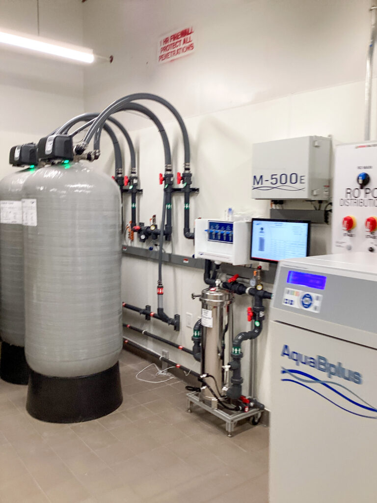 Industrial water filtration system featuring two large gray tanks, an M-500E control unit, a digital display, and an AquaPlus purification unit connected by piping.