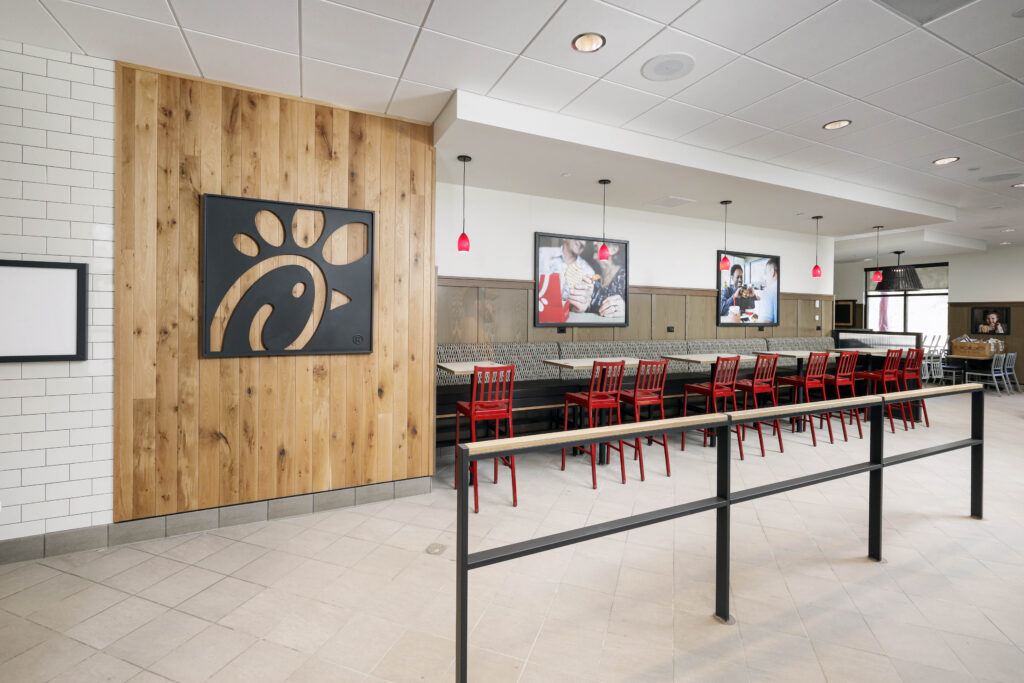 Interior of a Chick-fil-A restaurant with wooden wall paneling, a large black company logo, and rows of red chairs and tables.