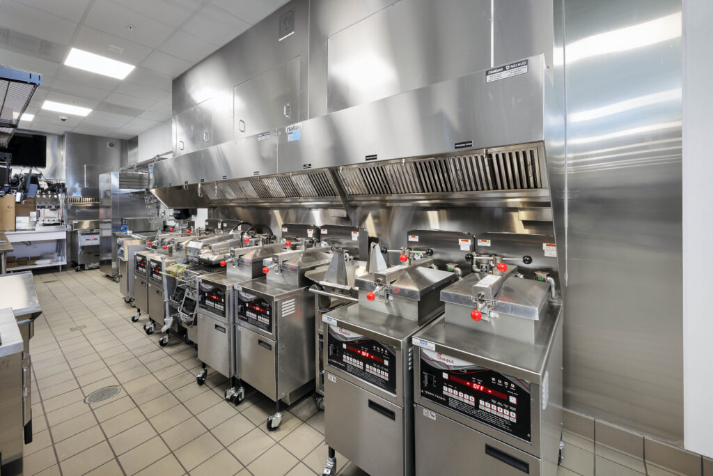 A row of commercial deep fryers under a stainless steel vent hood in a professional kitchen.