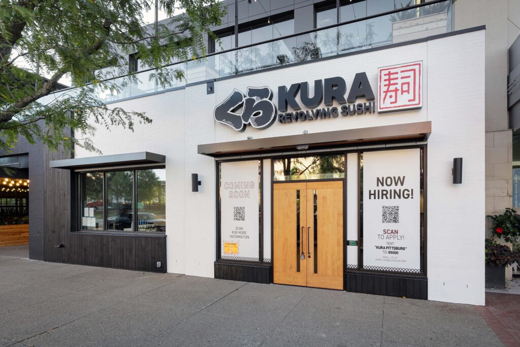 Exterior of a Kura Revolving Sushi restaurant with “Coming Soon” and “Now Hiring” signs displayed in the windows.