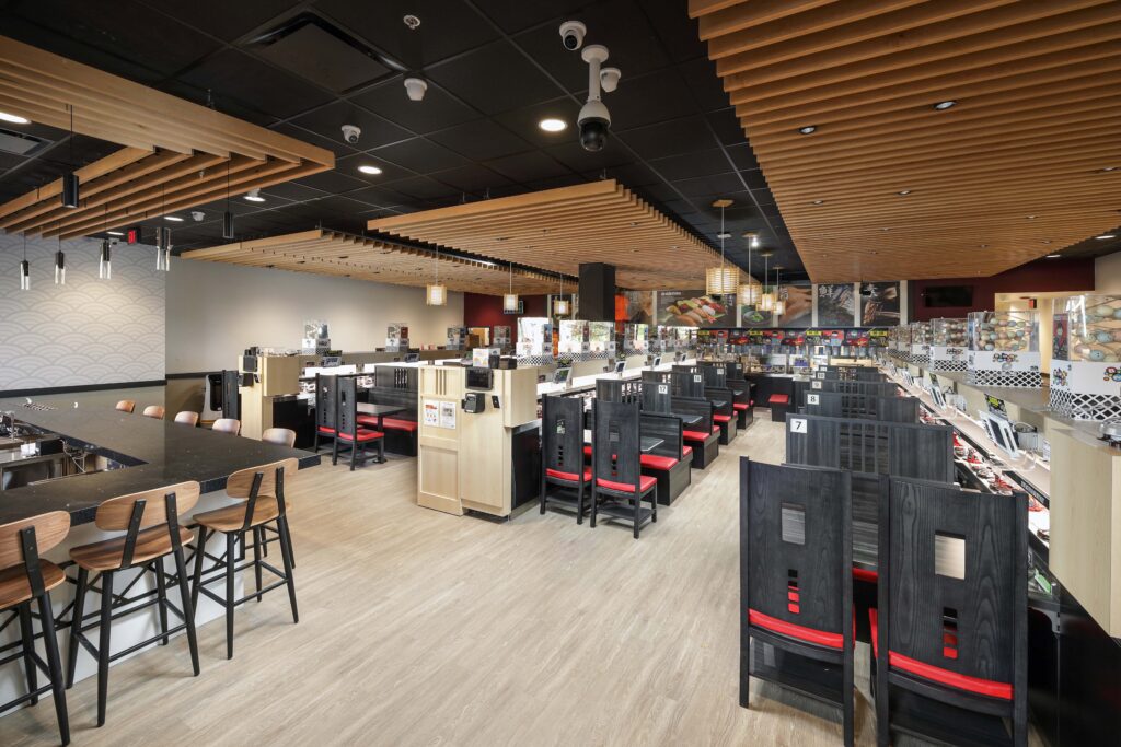 Interior of a sushi restaurant with wooden slatted ceilings, counter seating, and individual booths with black chairs and red cushions.