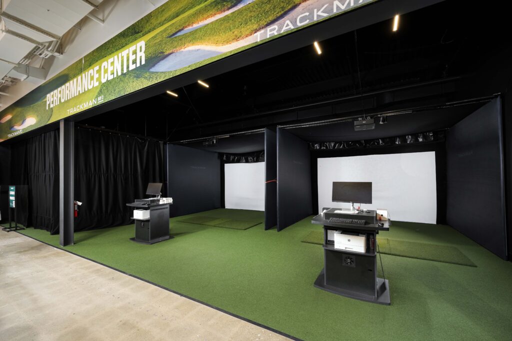 Two indoor golf simulator bays featuring green turf, large impact screens, and computer kiosks.