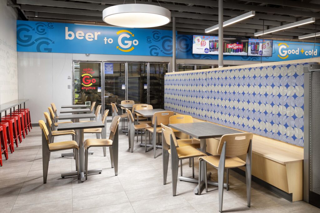 Dining area inside a convenience store with “beer to Go” signage above glass coolers and a row of tables and chairs.