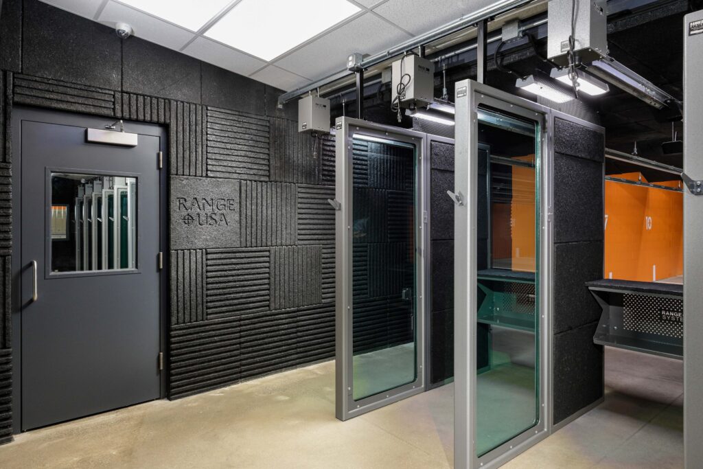 Interior of a shooting range featuring a door with a small window, acoustic wall paneling labeled “RANGE USA,” and several glass-enclosed shooting stalls.