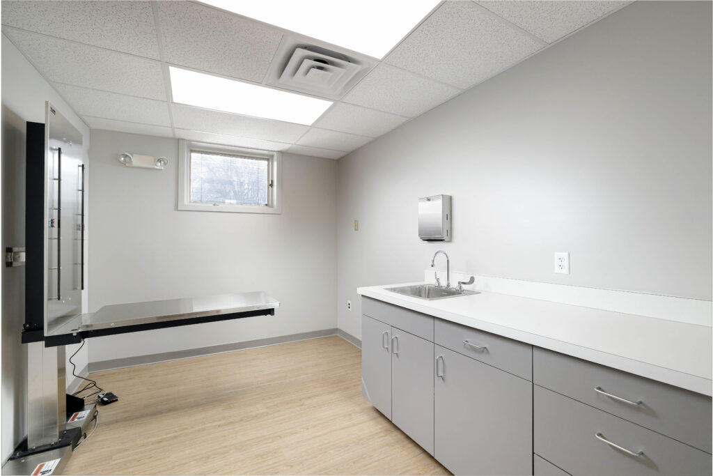 A medical exam room featuring a stainless steel wall-mounted exam table, a sink with cabinetry, and a small window.