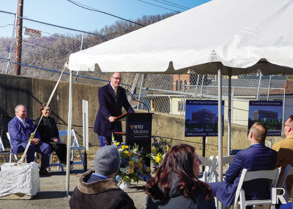 A man in a suit speaks at a podium during a groundbreaking ceremony for the St. Joseph Regional Cancer Center, with renderings of the future building displayed on easels.
