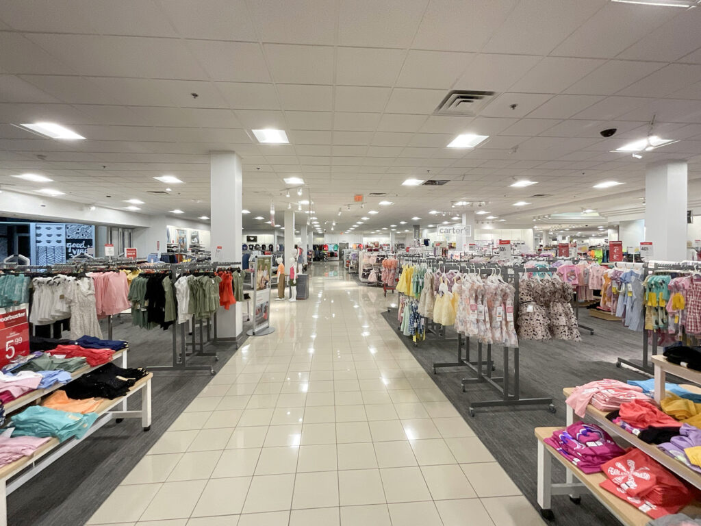 Wide aisle in a clothing store lined with racks of children’s apparel and display tables with folded garments.