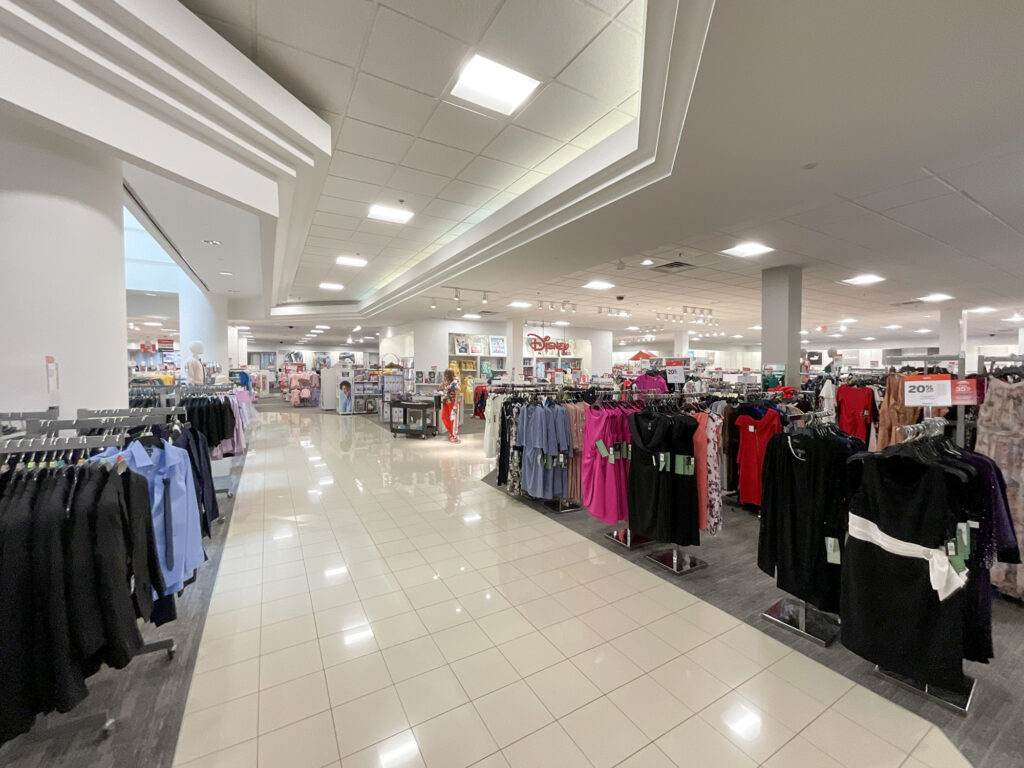 Inside a clothing department store with rows of garment racks and a tile floor.