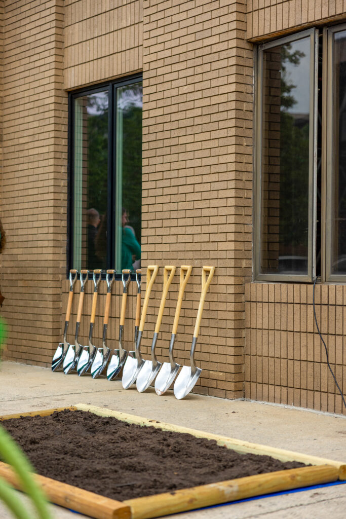 Seven ceremonial shovels leaned against a brick building beside a rectangular patch of soil.