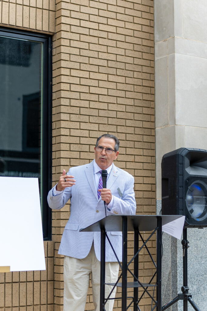 A man in a light-colored blazer and tie speaks into a microphone at a lectern.