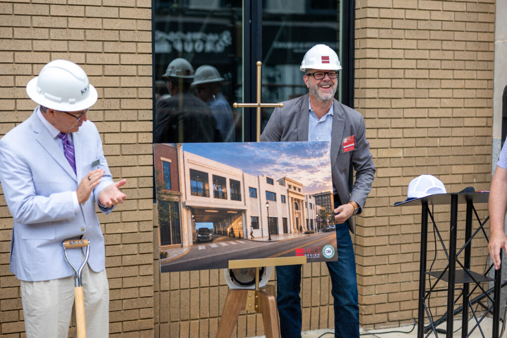 Two men in hard hats stand before a brick building; one holds a framed architectural rendering of a multi-story building facade and street entrance.