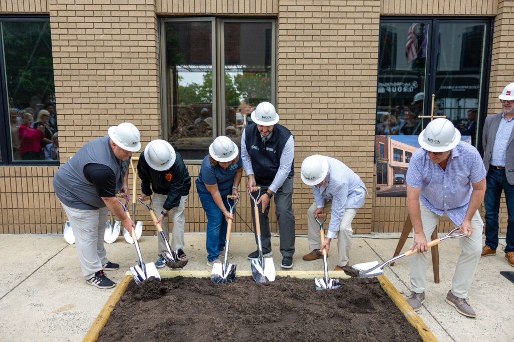 Six people wearing white hard hats ceremonially break ground with shovels in a wooden planter box in front of a brick building.