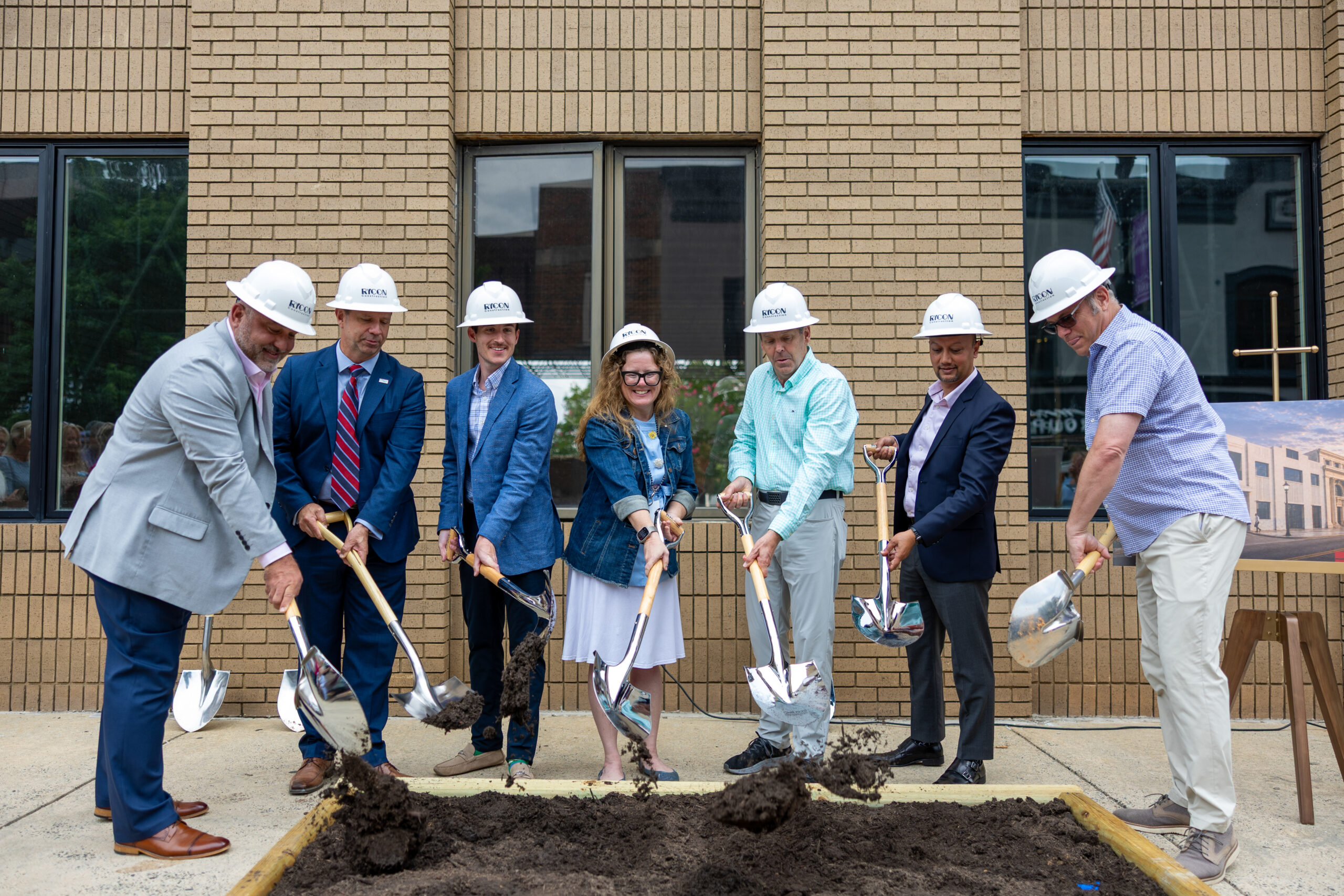 Seven people in hard hats participate in a ceremonial groundbreaking with shovels in front of a brick building.