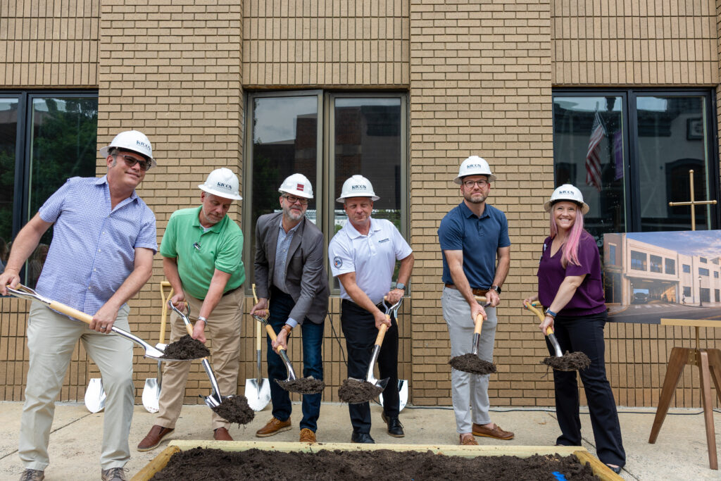 Six people in hard hats participate in a ceremonial groundbreaking with shovels in front of a brick building.