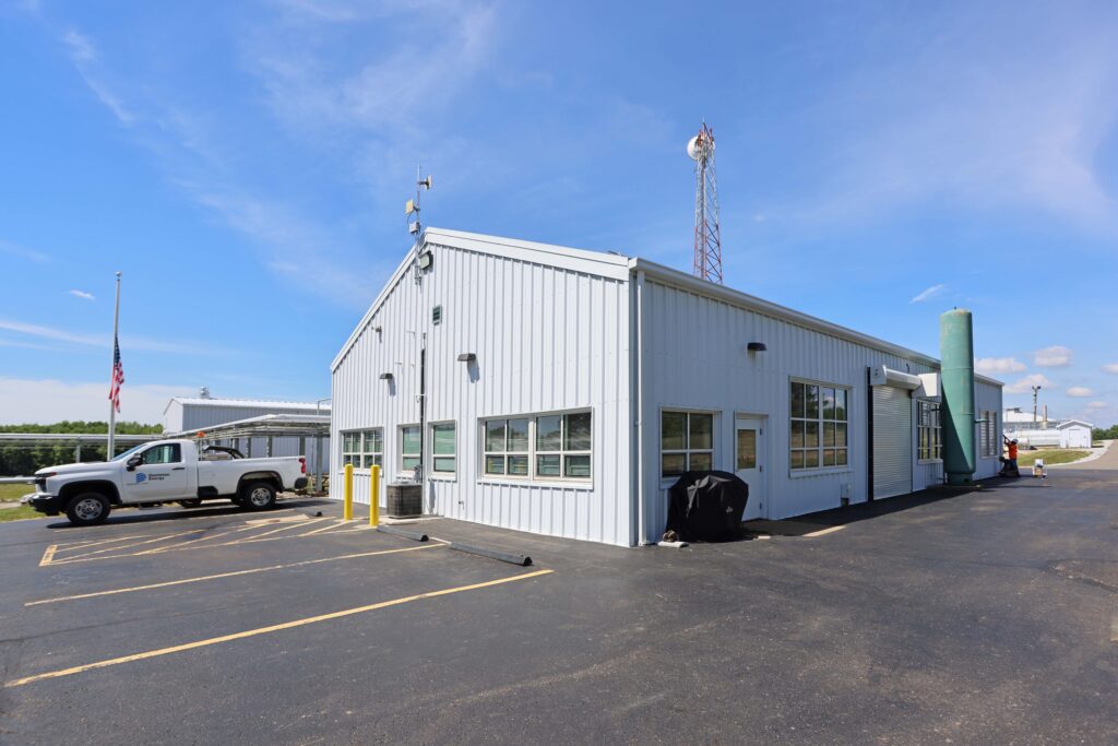 A white metal utility building with a parking lot, a pickup truck, and a tall antenna tower under a clear blue sky.