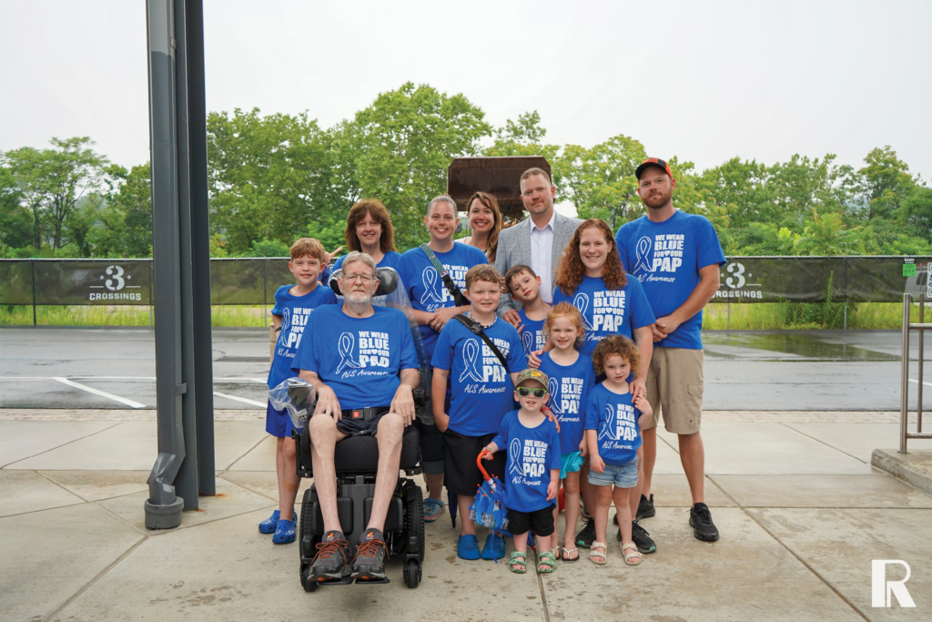 A group of people, including a man in a wheelchair, wearing matching blue “We Wear Blue for Our Pap” ALS awareness t-shirts while posing for a photo outdoors.