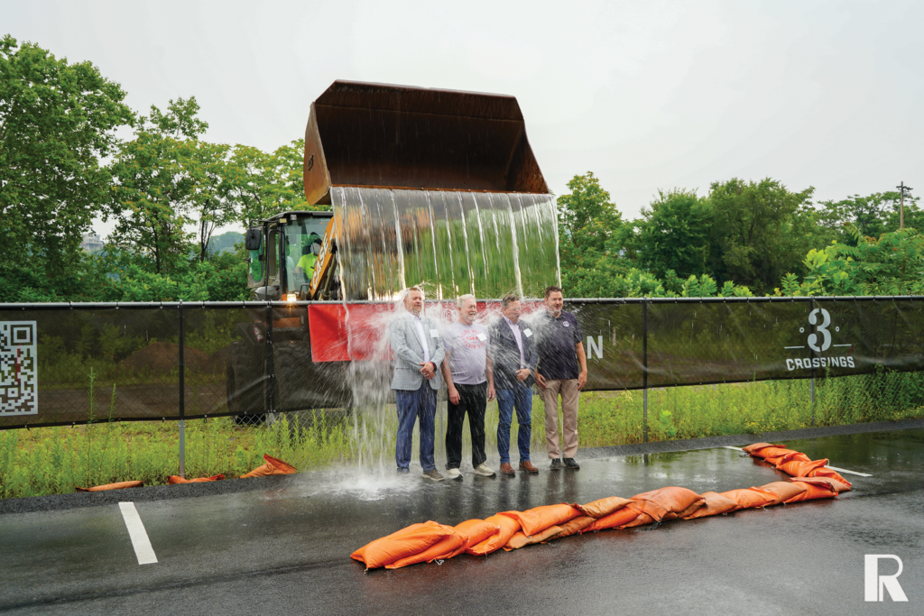 Four men standing in front of a construction fence getting drenched by water pouring from a front-end loader bucket.