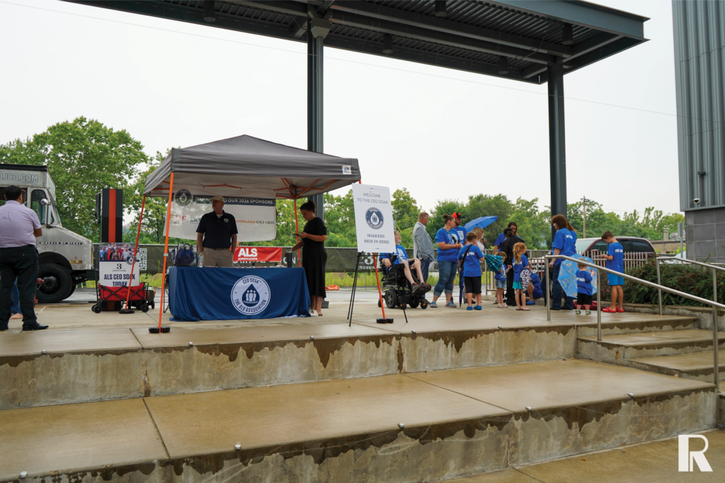 A canopy tent at an ALS Association “CEO Soak” fundraising event with volunteers and attendees gathered on an outdoor concrete stage.