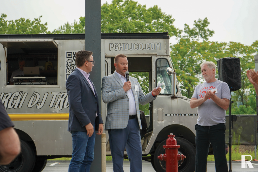 A man in a plaid blazer speaks into a microphone, standing next to another man in a blazer and a man in a t-shirt in front of a food truck.