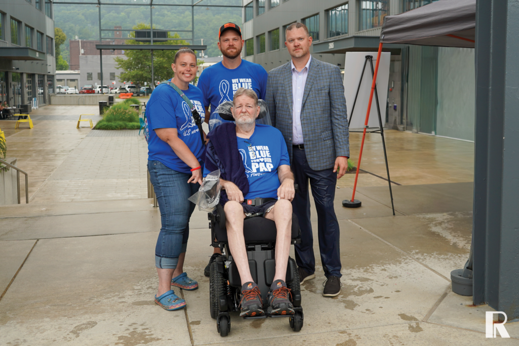 Four people posing for a photo outdoors, including a man in a wheelchair wearing a “We Wear Blue for Our Pap” t-shirt.