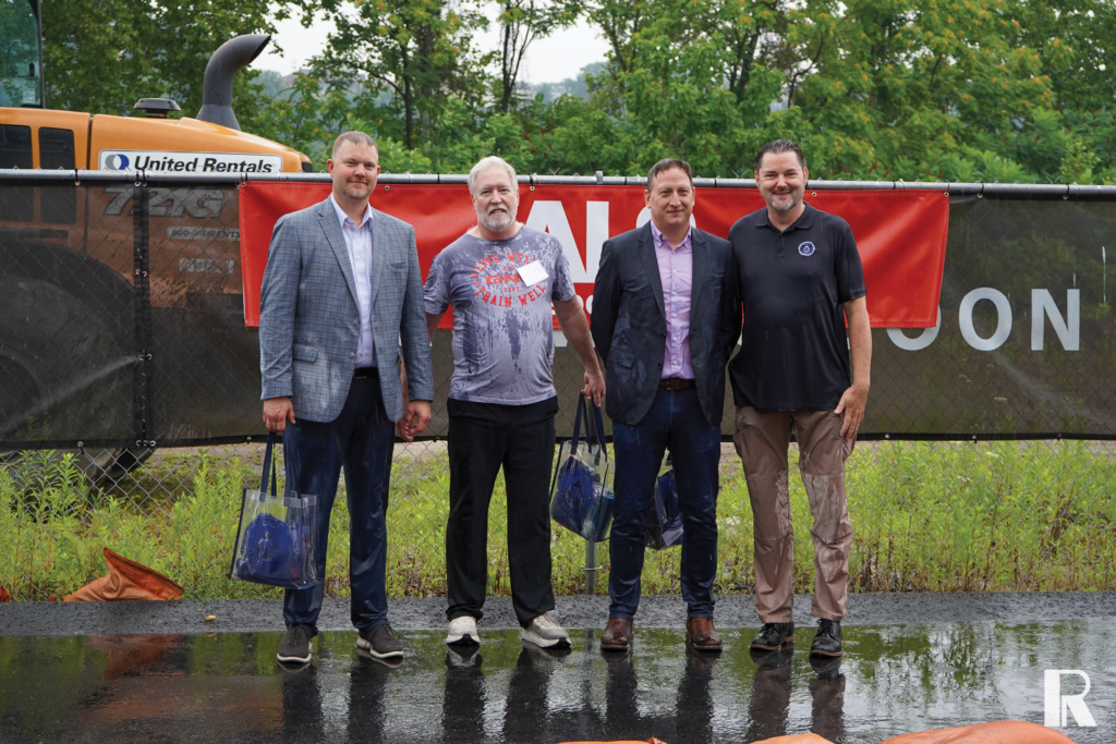 Four men stand in a row outdoors in front of a chain-link fence and a construction vehicle.